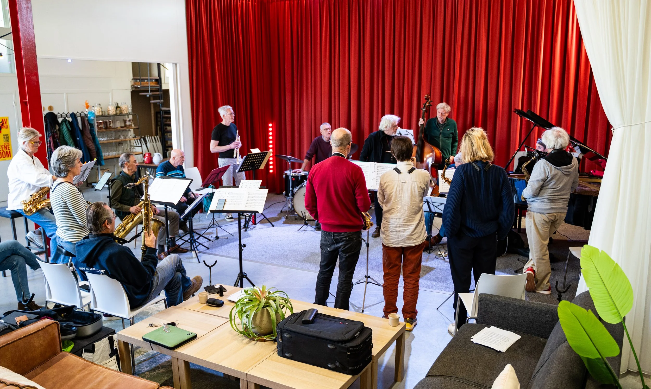 Een muzikale repetitie met verschillende muzikanten in een zaal met een rood gordijn op de achtergrond en zitplaatsen rondom. Muzikanten spelen saxofoon, viool, piano, een stokken of -een trommel, en een dubbelbas.