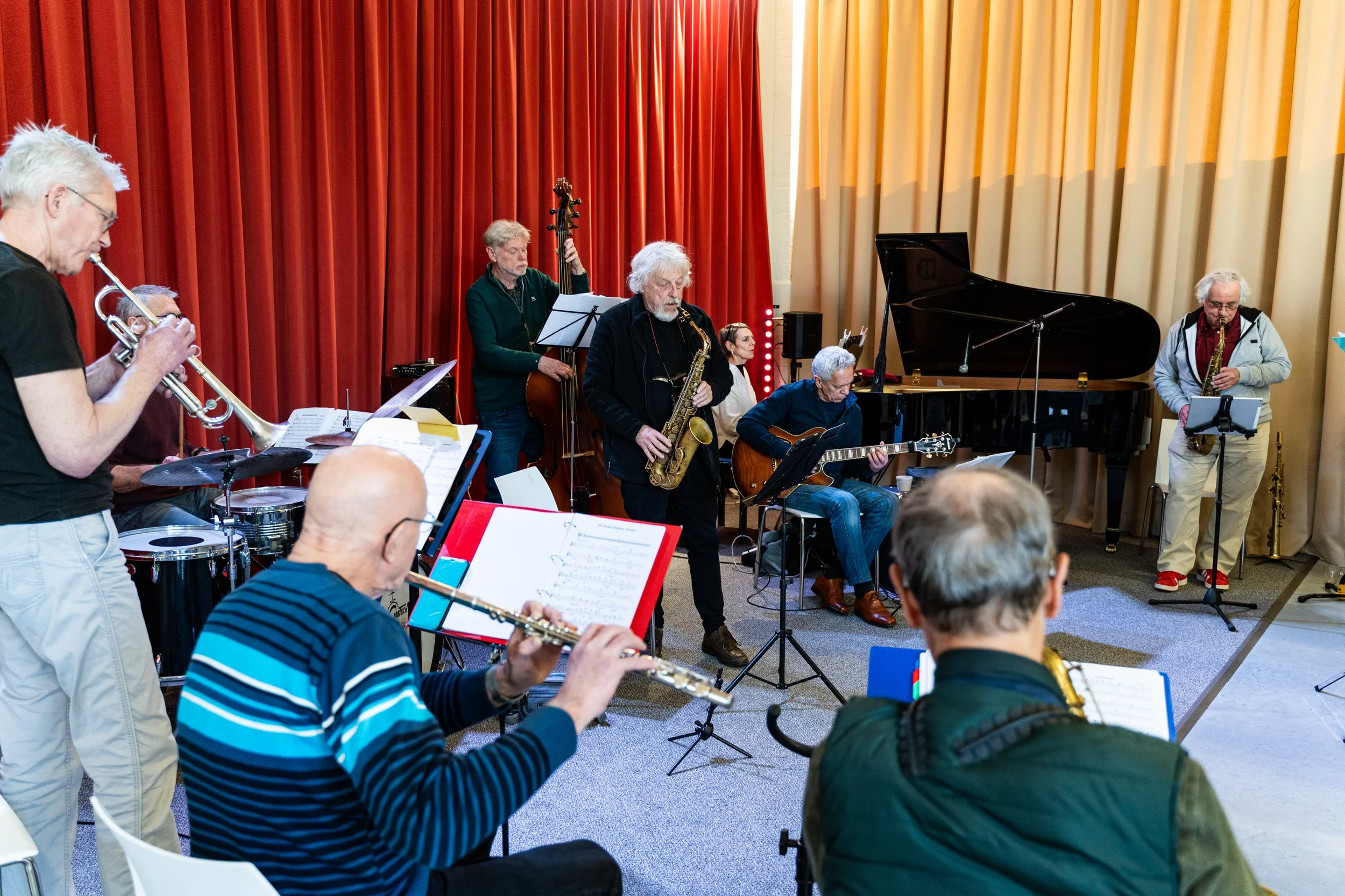 Mannen en vrouwen spelen jazzmuziek in een performanceruimte met rode en beige gordijnen, inclusief een piano en blaasinstrumenten.