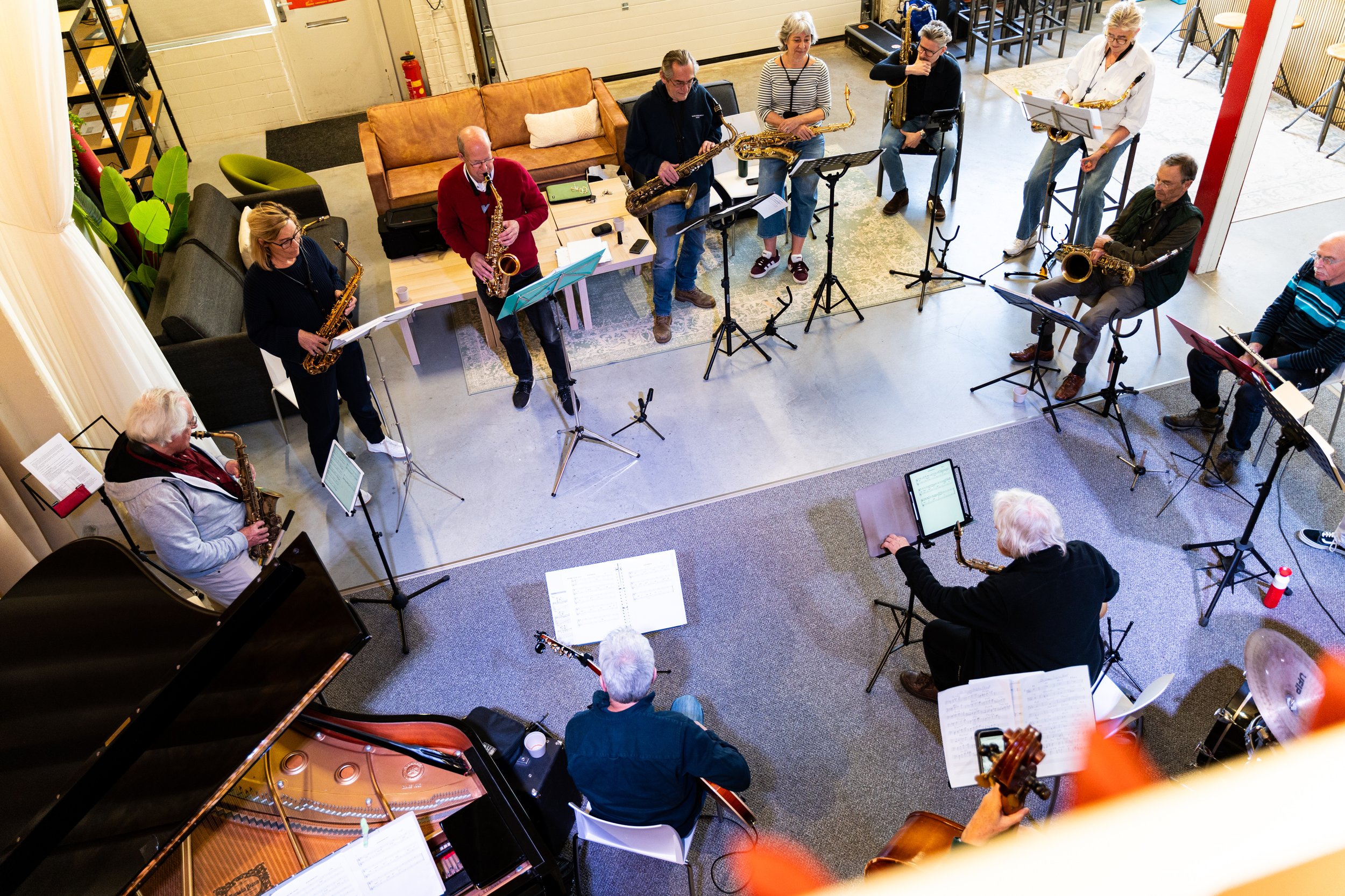 Muziekband die repeteert in een zaal, bestaande uit verschillende muzikanten met instrumenten zoals saxofoon, piano en basgitaar.