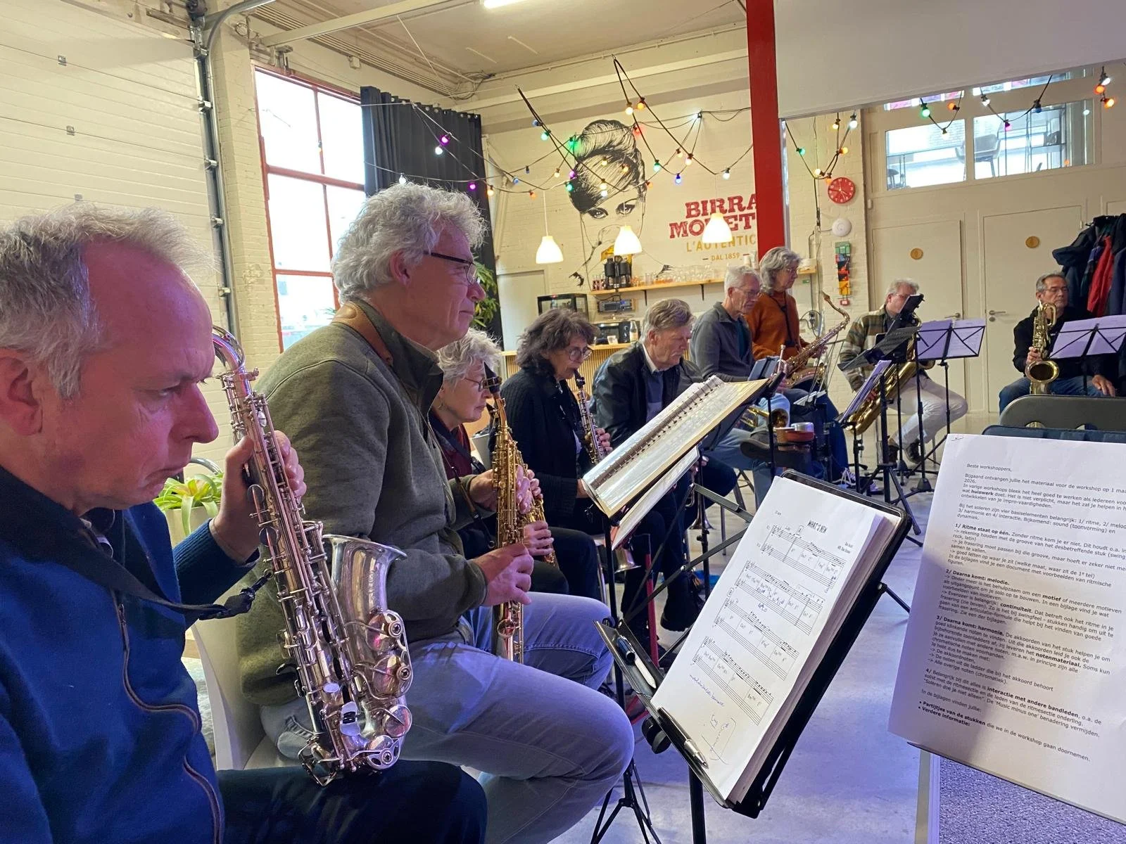 Een groep oudere muzikanten die samen in een repetitieruimte spelen, met bladmuziek op standaard en een muur met een grote posters met een vrouw en tekst in het Frans.