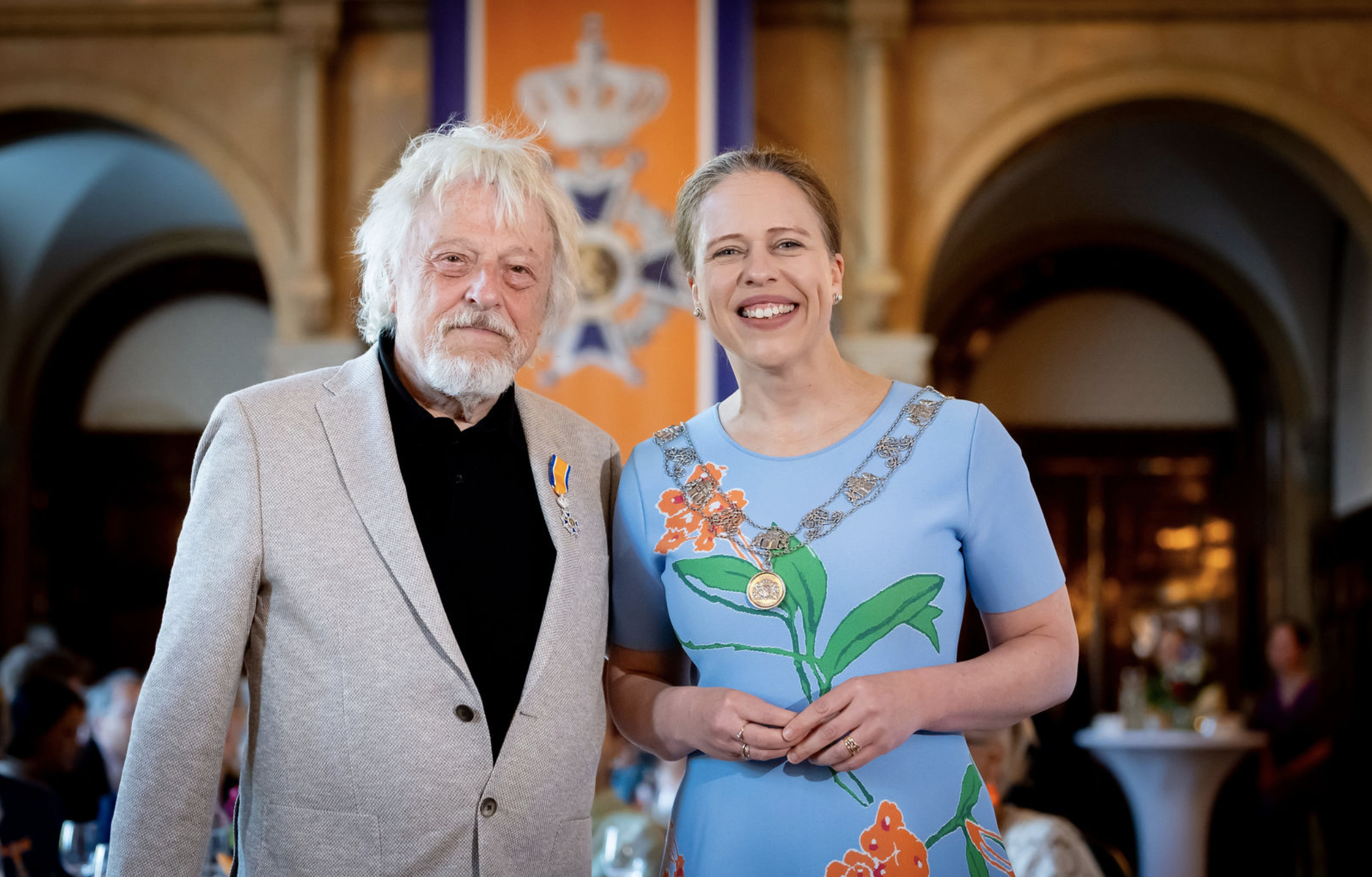 Man en vrouw poseren in een feestzaal, met een vlag en embleem op de achtergrond, lachend en in formele kleding.