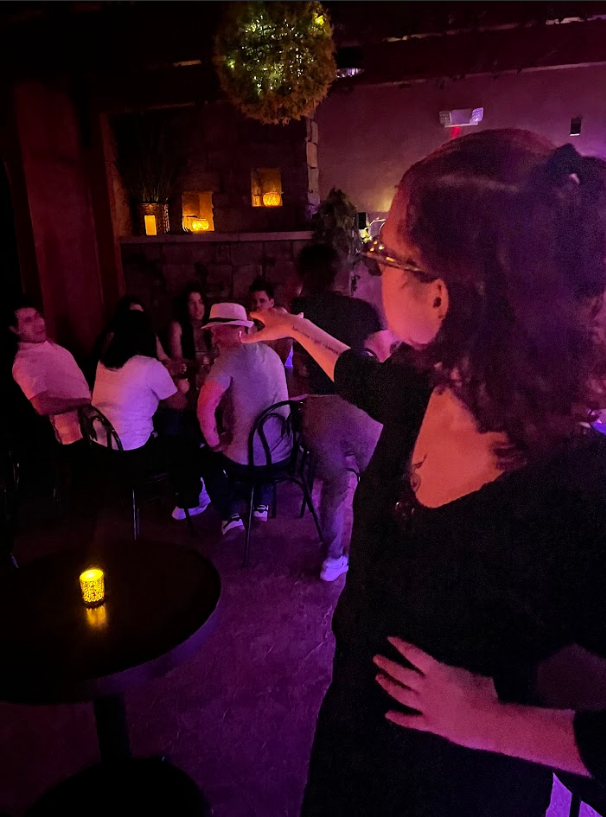 A woman with glasses and curly hair stands with her hand on her hip and points at a group of people sitting at a table in a dimly lit, cozy bar or restaurant with warm lighting and decorative plants.