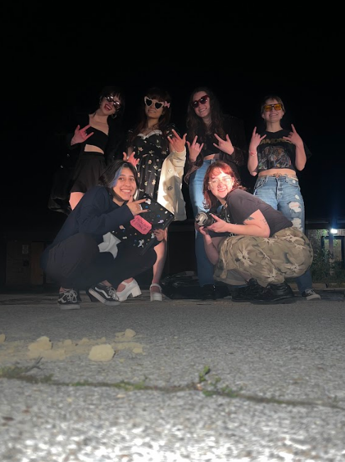 Five young women posing together outdoors at night, some standing and some crouching, all smiling and making peace signs or hand gestures. They are dressed casually, with sunglasses and streetwear, against a dark background with some lights.