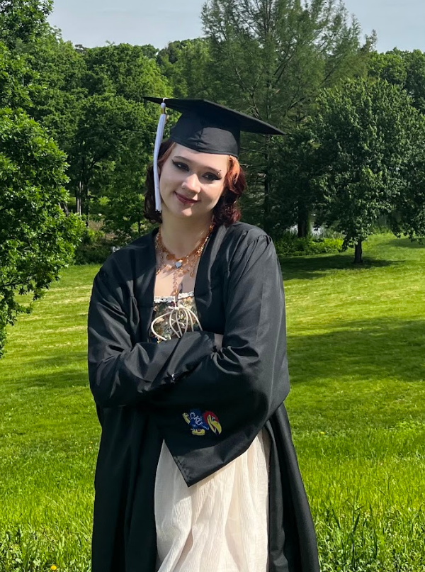 A young woman in a graduation cap and gown standing outdoors on a sunny day, with green trees and a grassy field in the background.