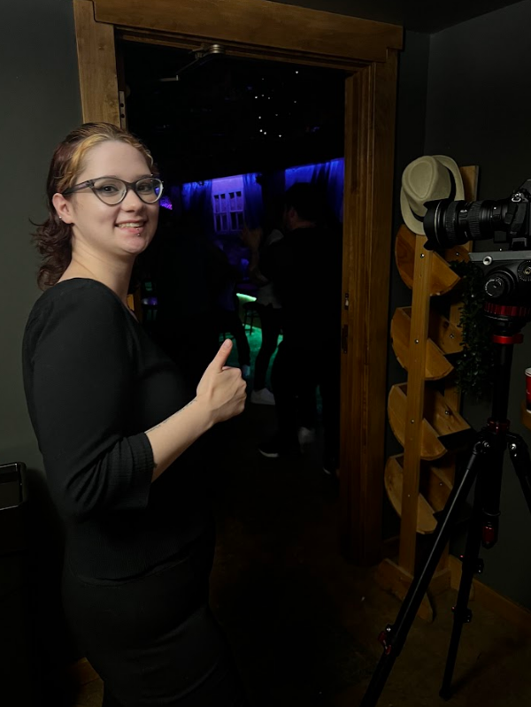 Woman with glasses, wearing a black top, smiling and pointing at a window with a view of an indoor space with people and blue and green lights.