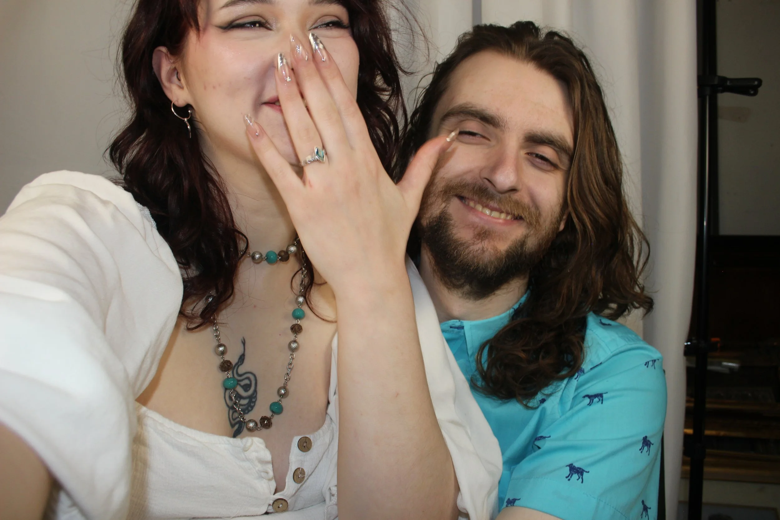 Two people, a woman with dark red hair and a man with long brown hair and beard, are smiling and appear to be sharing a joyous moment. The woman covers her mouth with her hand, showing polished nails and jewelry, while the man grins happily.