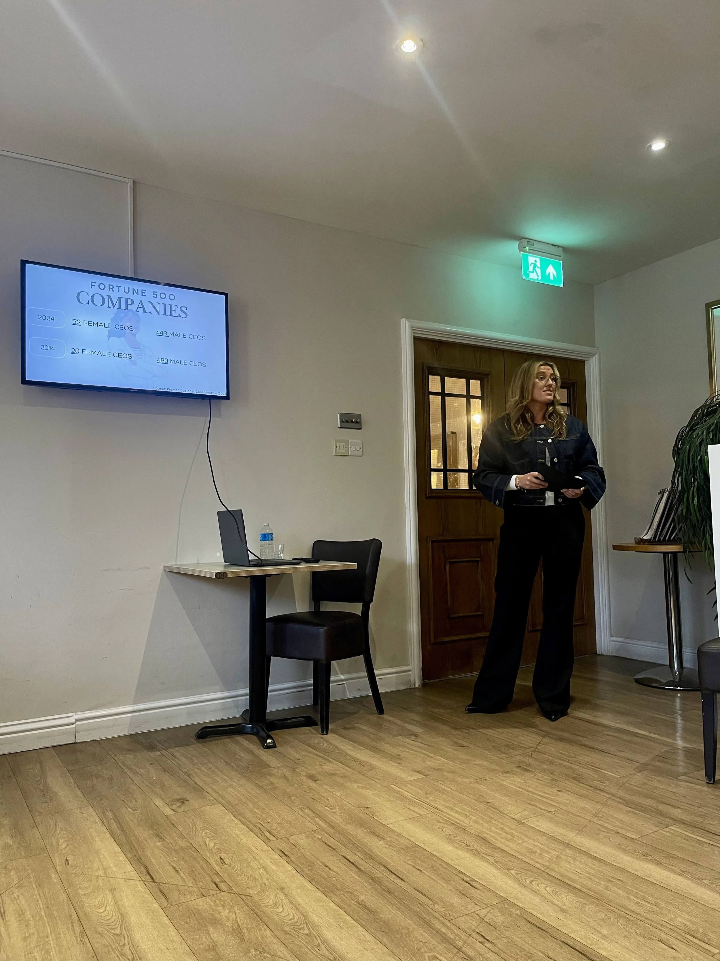 A woman in business attire giving a presentation in a conference room with a wooden floor, a mounted screen displaying company statistics, and an emergency exit sign above a door.