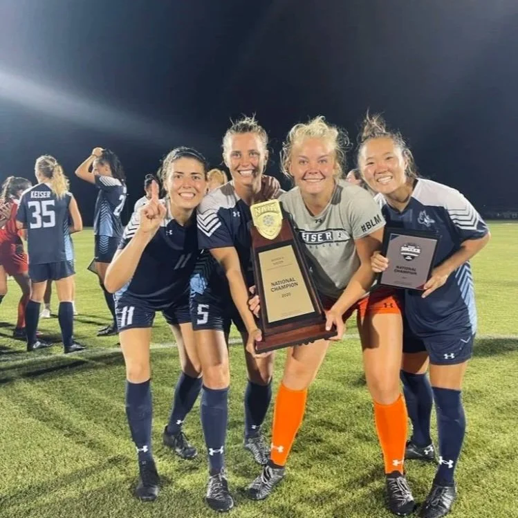 Four women soccer players celebrate with a championship plaque on a soccer field at night. They are smiling and posing for the camera, with two women holding the plaque and one woman pointing upwards.