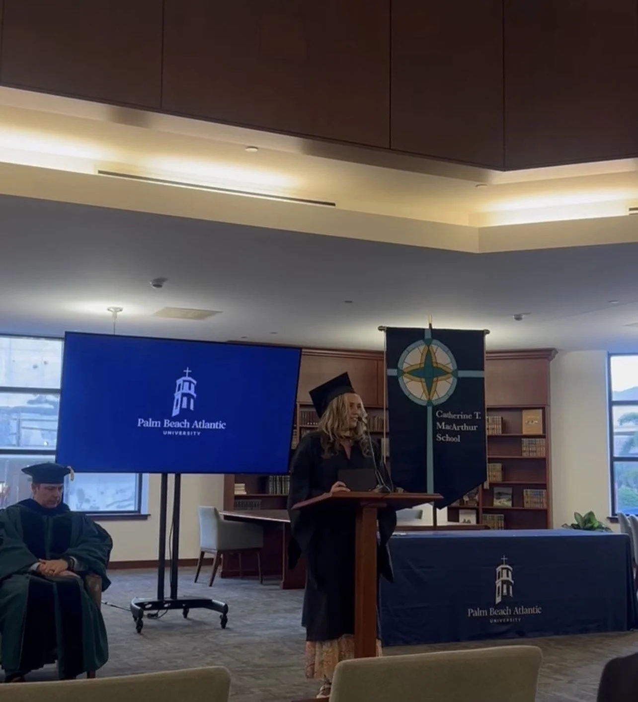 A graduation ceremony at Palm Beach Atlantic University with a woman in a graduation gown and cap speaking at a podium, and a man in academic regalia seated nearby. There are screens and banners displaying the university's logo and name.