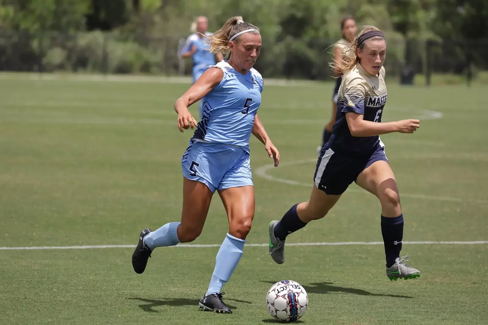 Two women are playing soccer on a grass field, one in a blue uniform and the other in a black uniform, with the woman in black kicking the ball.
