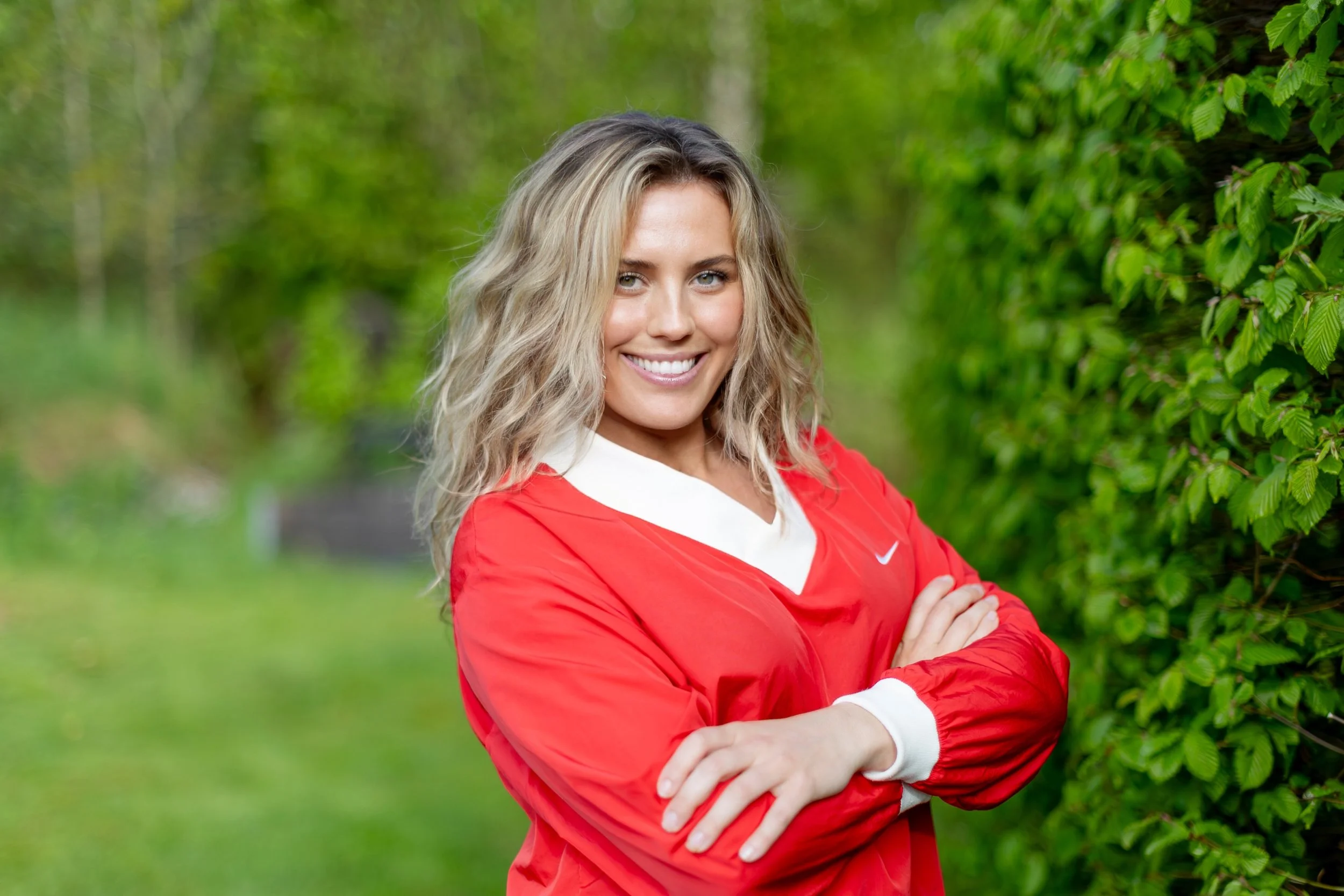 A smiling woman with blonde hair wearing a red sports jacket standing outdoors among green foliage.