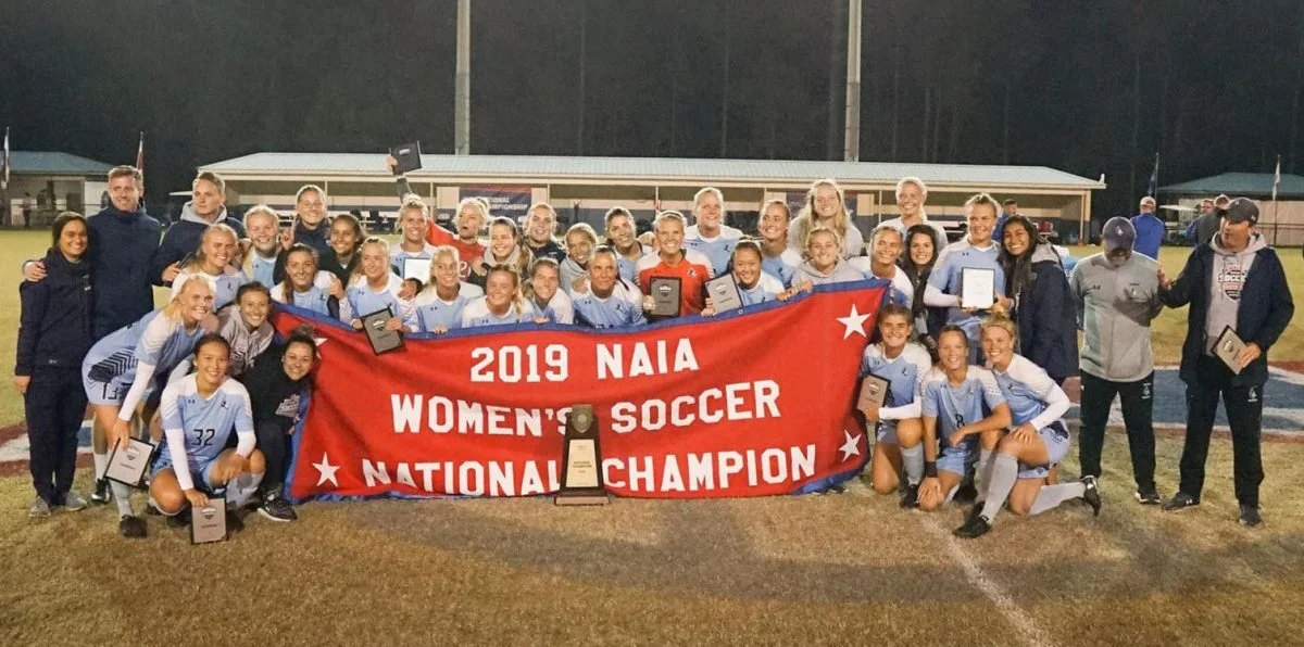 Women’s soccer team holding a red banner that reads '2019 NAIA Women’s Soccer National Champion,' with trophies and awards, on a soccer field at night.