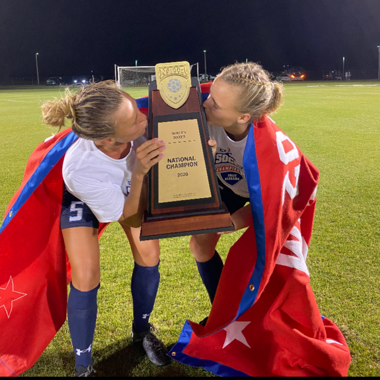 Two female soccer players kissing and holding a trophy after winning a game, wearing white jerseys, navy shorts, and wrapped in red flags with white stars and blue trim, on a soccer field at night.