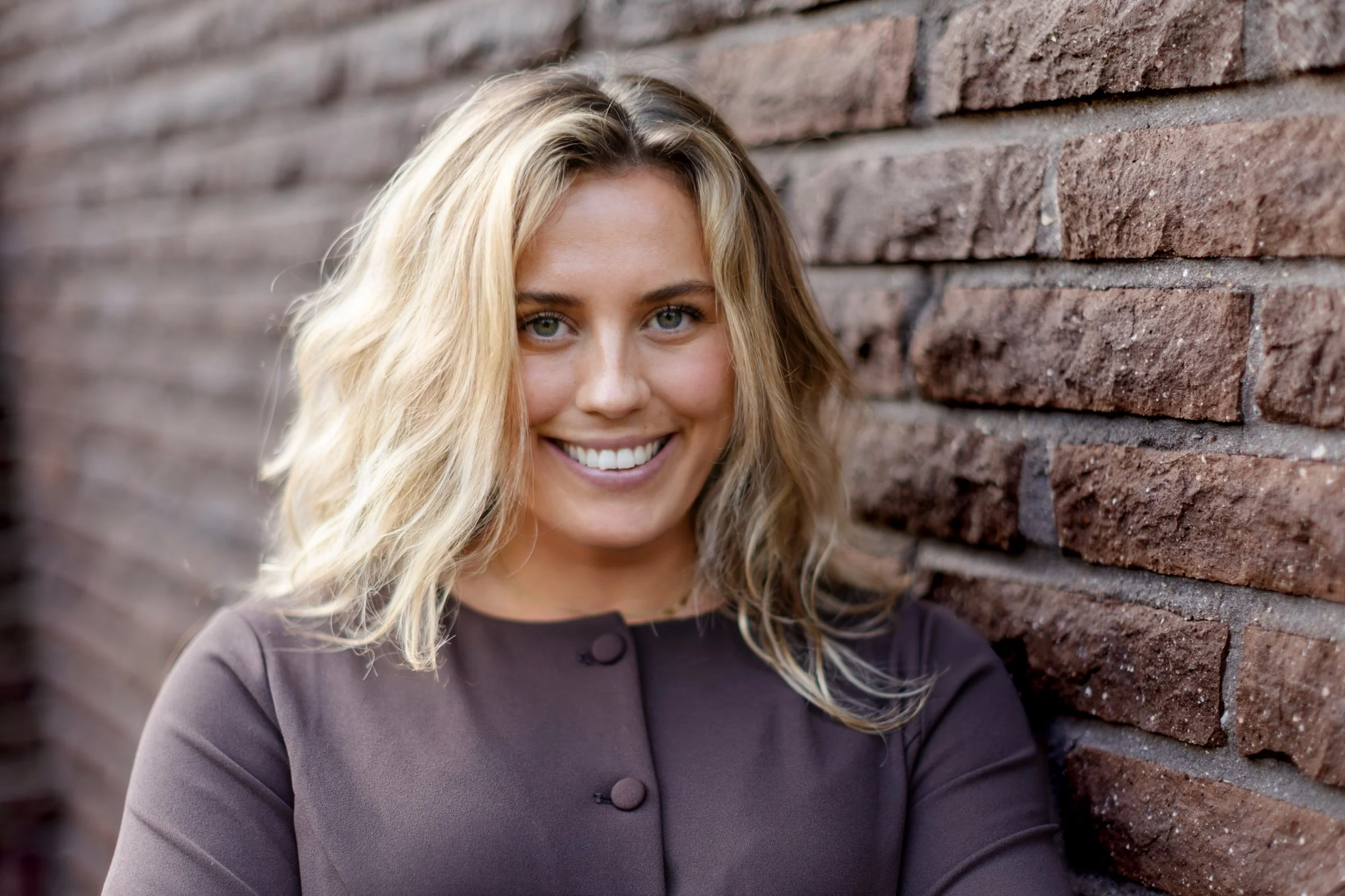 A woman with long blonde hair and blue eyes smiling, wearing a dark top, leaning against a brick wall.
