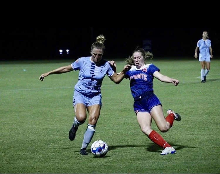 Two female soccer players competing for the ball on a field at night, with a third player in the background.
