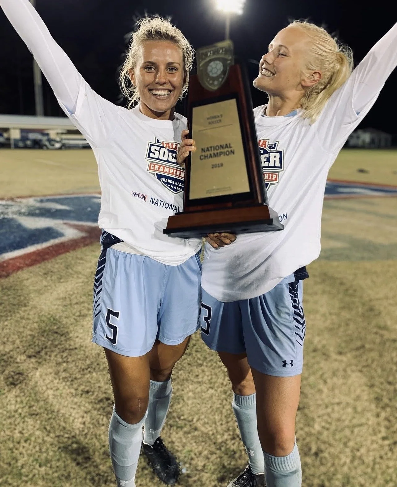 Two female soccer players celebrating on the field, holding a trophy that reads "National Champion." They are wearing white jerseys and light blue shorts, with bright smiles on their faces at night.