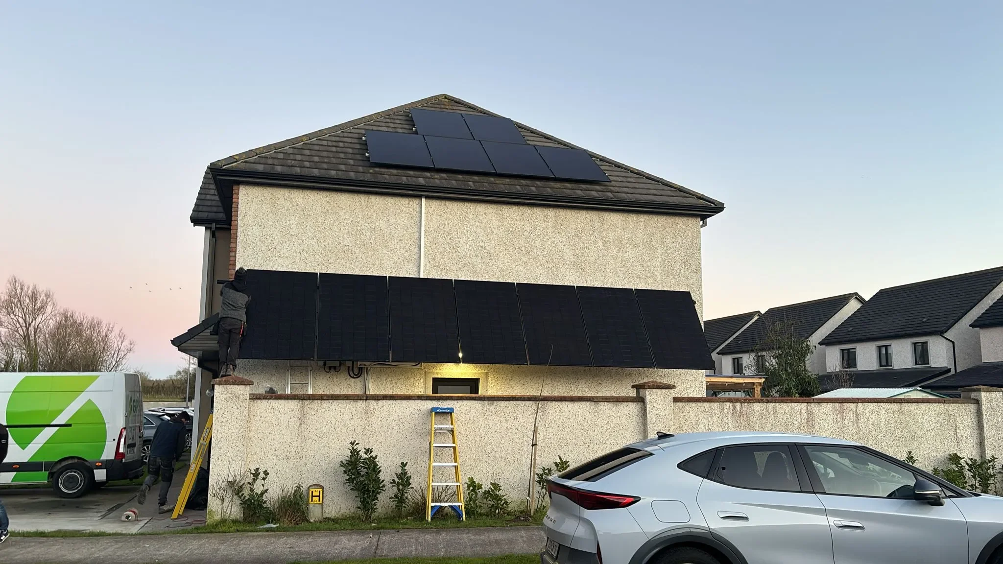 A two-story house with a person installing black solar panels on the roof, which already has several solar panels. There are parked cars in the front driveway, and two ladders leaning against the house. The sky is clear and the surroundings include other residential houses and some trees.
