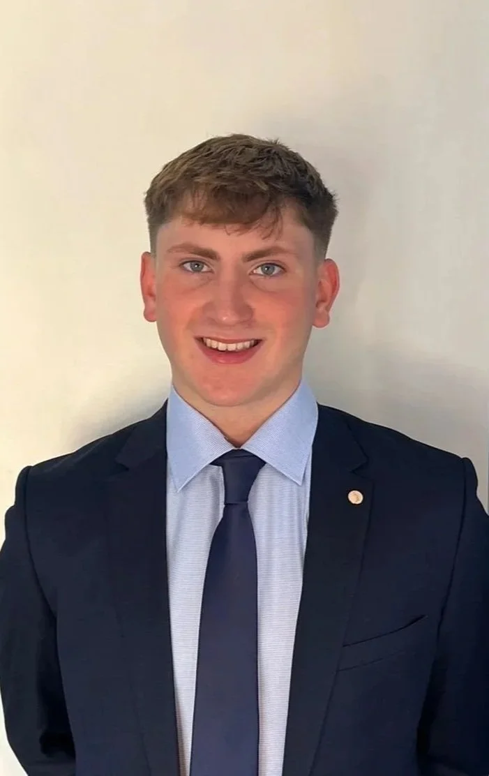 A young man with short brown hair, blue eyes, and fair skin, wearing a dark suit, light blue dress shirt, dark blue tie, standing against a plain light-colored wall, smiling.
