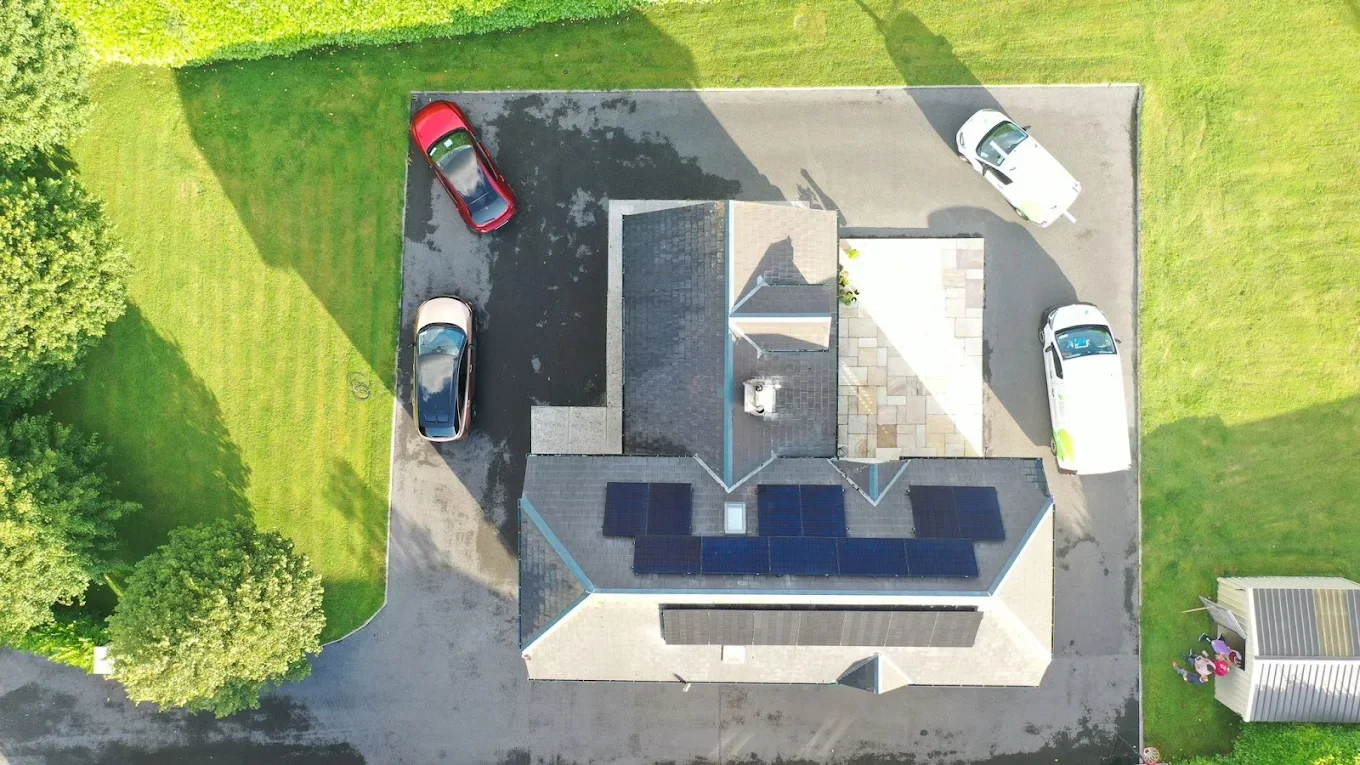 An aerial view of a house with a dark roof equipped with solar panels, surrounded by a parking area with several parked cars, green grass, and trees.