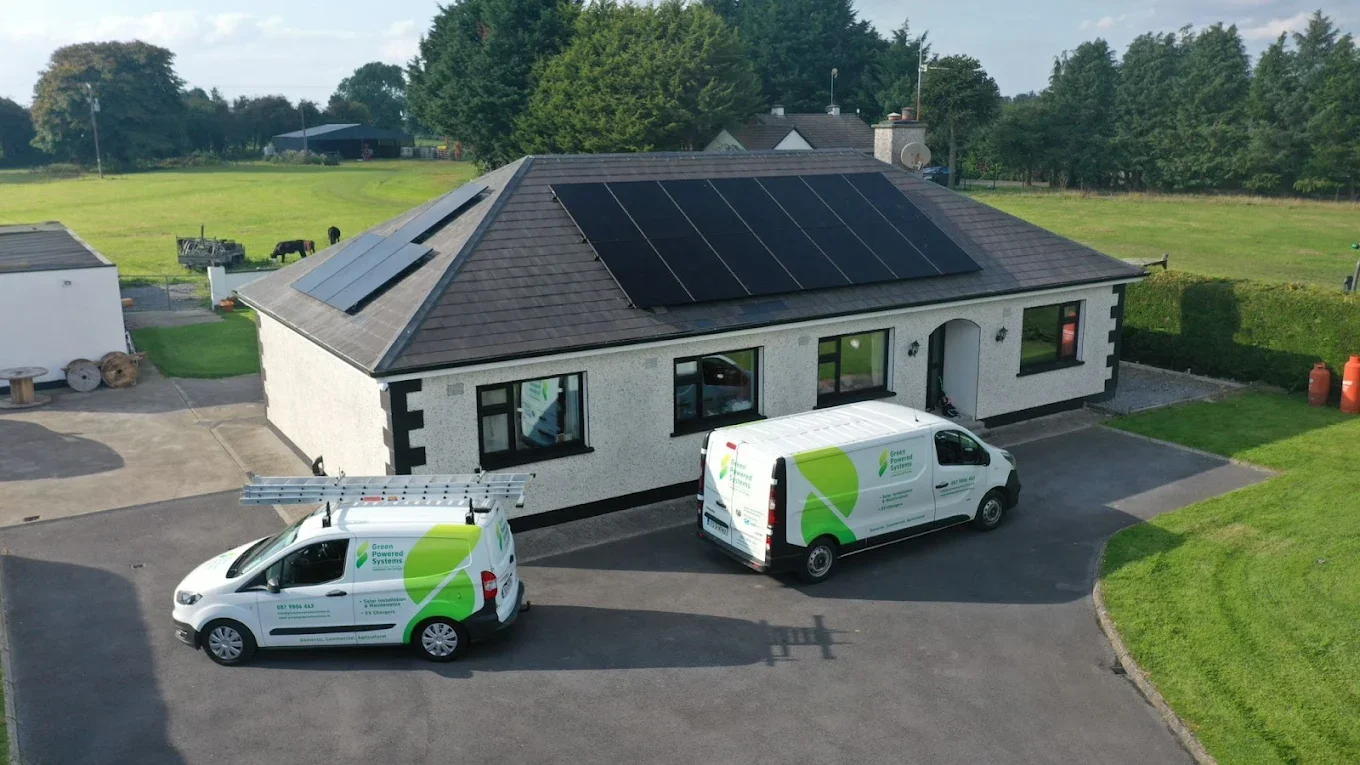 Two white service vans with green branding parked in front of a house with solar panels on the roof. The house has a driveway and surrounding green lawns.