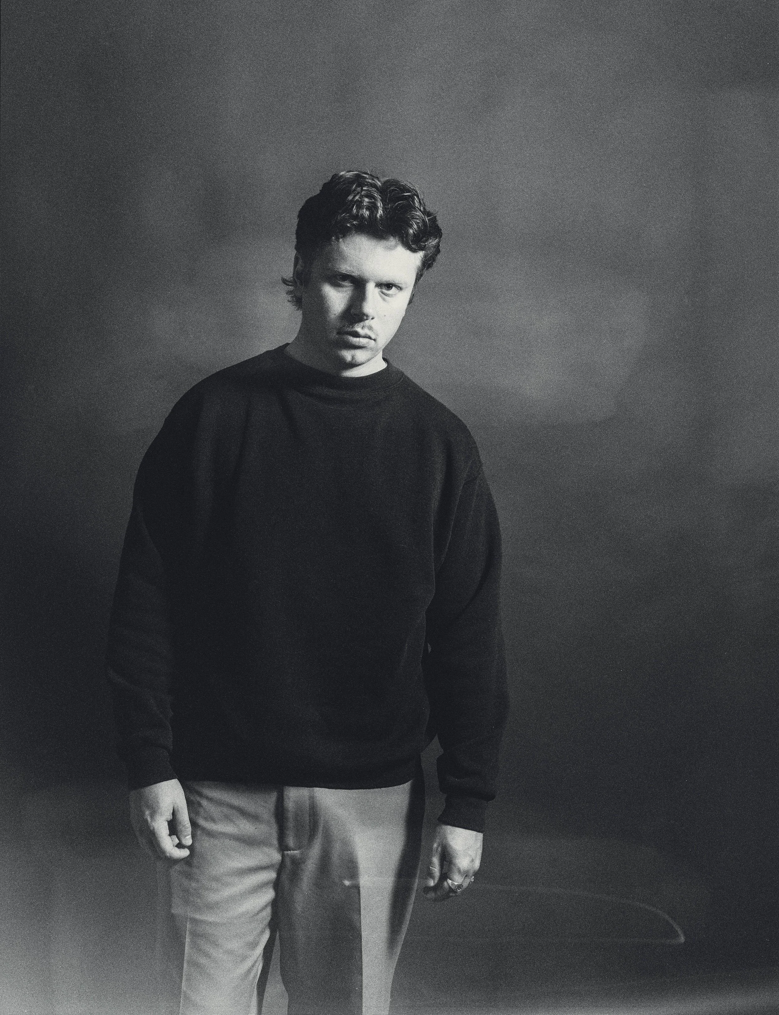 Schwarz-weiß-Foto eines jungen Mannes mit dunklen, gewellten Haaren, trug einen schwarzen Pullover und helle Hosen, steht vor einem dunklen Hintergrund.