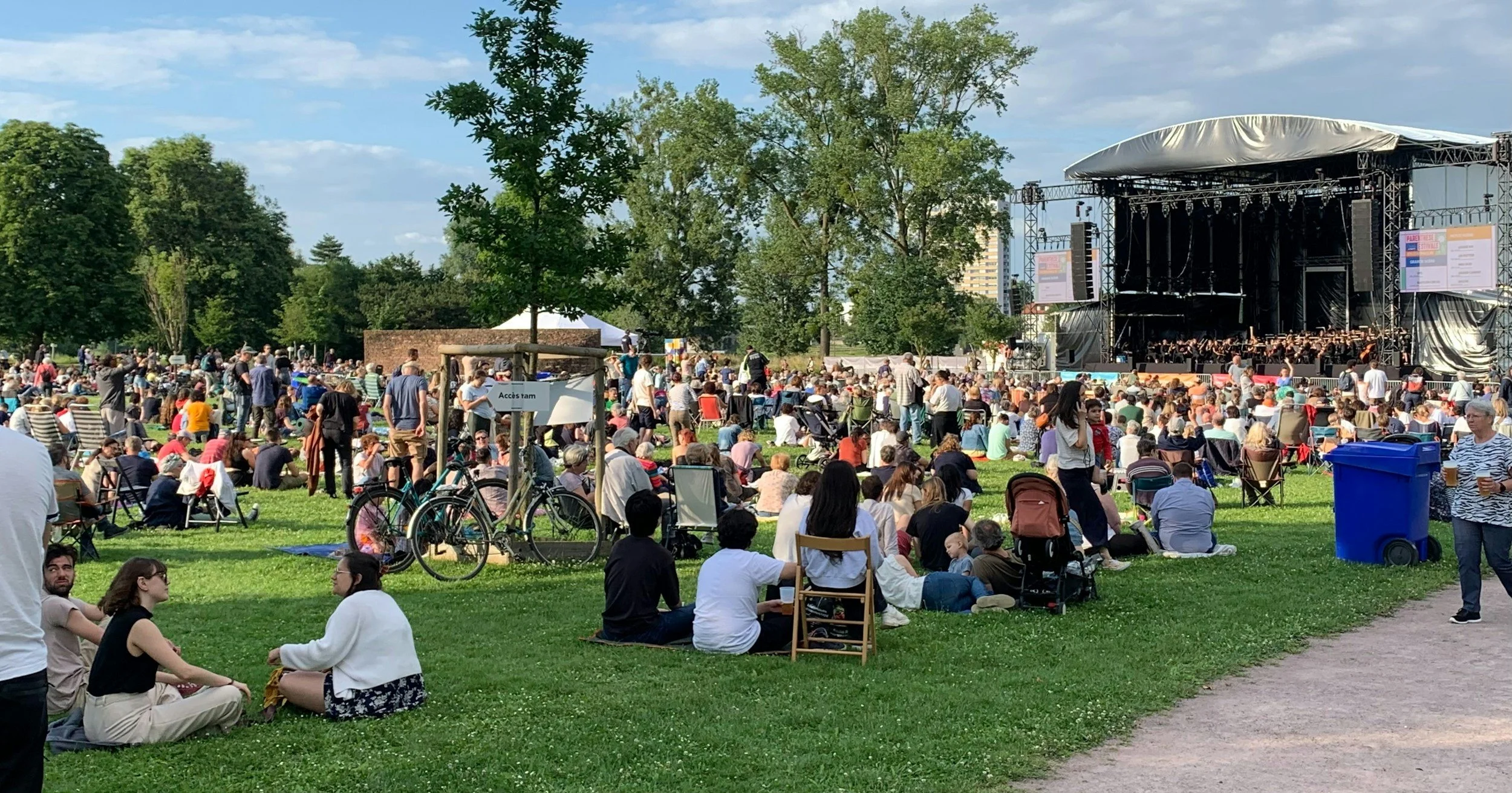 People attending an outdoor concert in a park with a large stage and orchesta, surrounded by trees and an open sky.