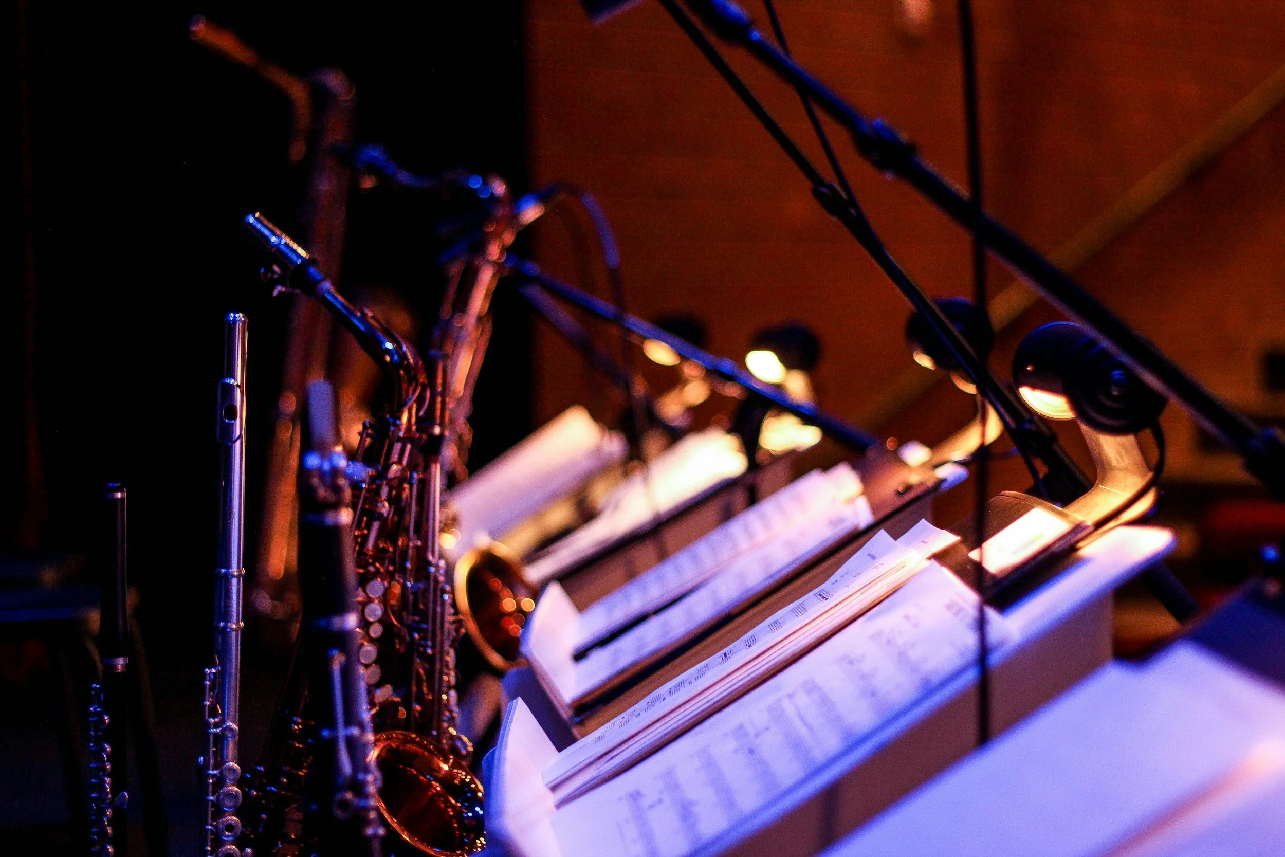 A row of music stands with sheet music and a collection of wind instruments, including clarinets and saxophones, illuminated by stage lighting.