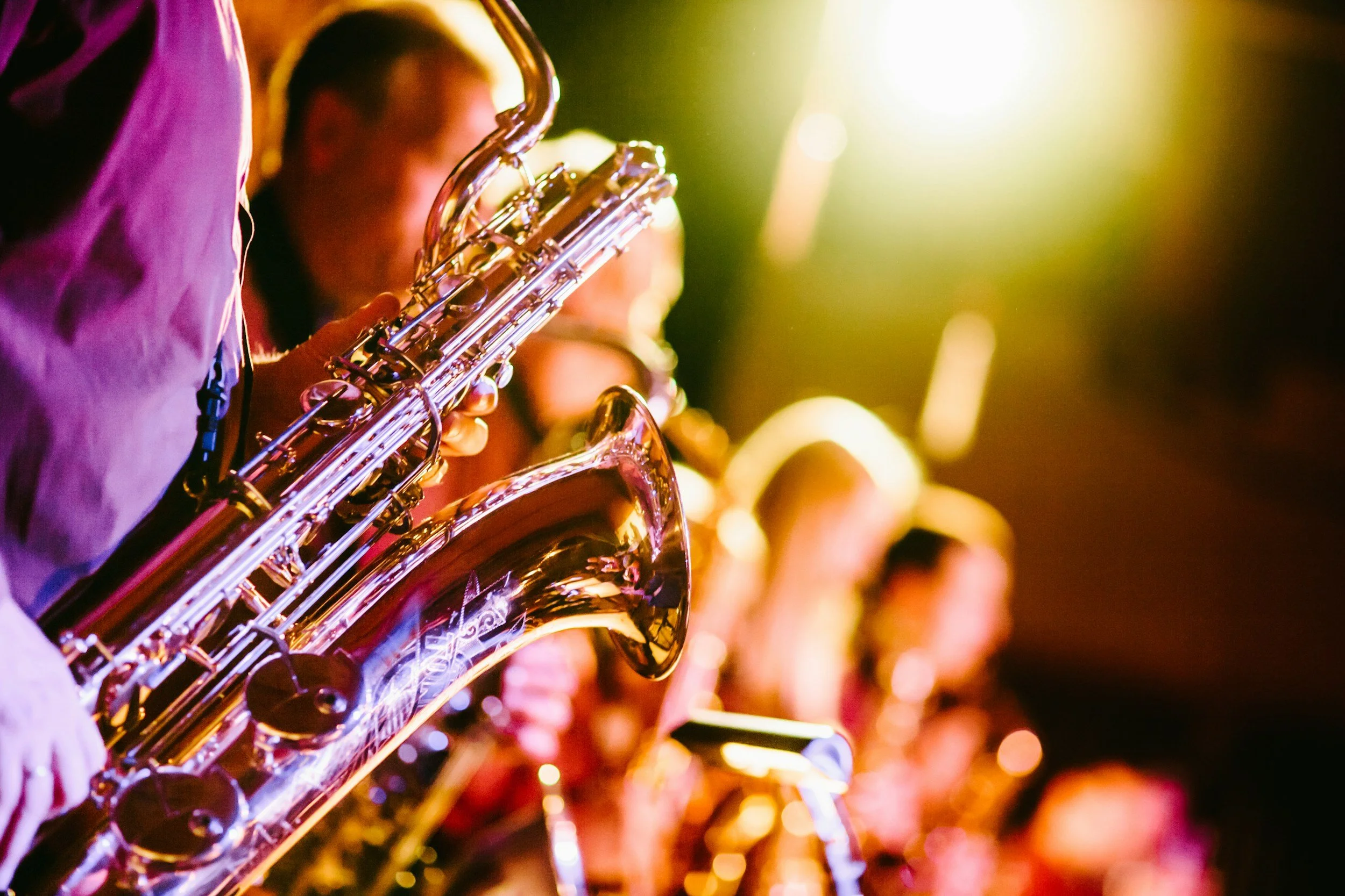 Close-up of a person playing a saxophone in a musical performance, with blurred musicians in the background and stage lighting creating a vibrant atmosphere.