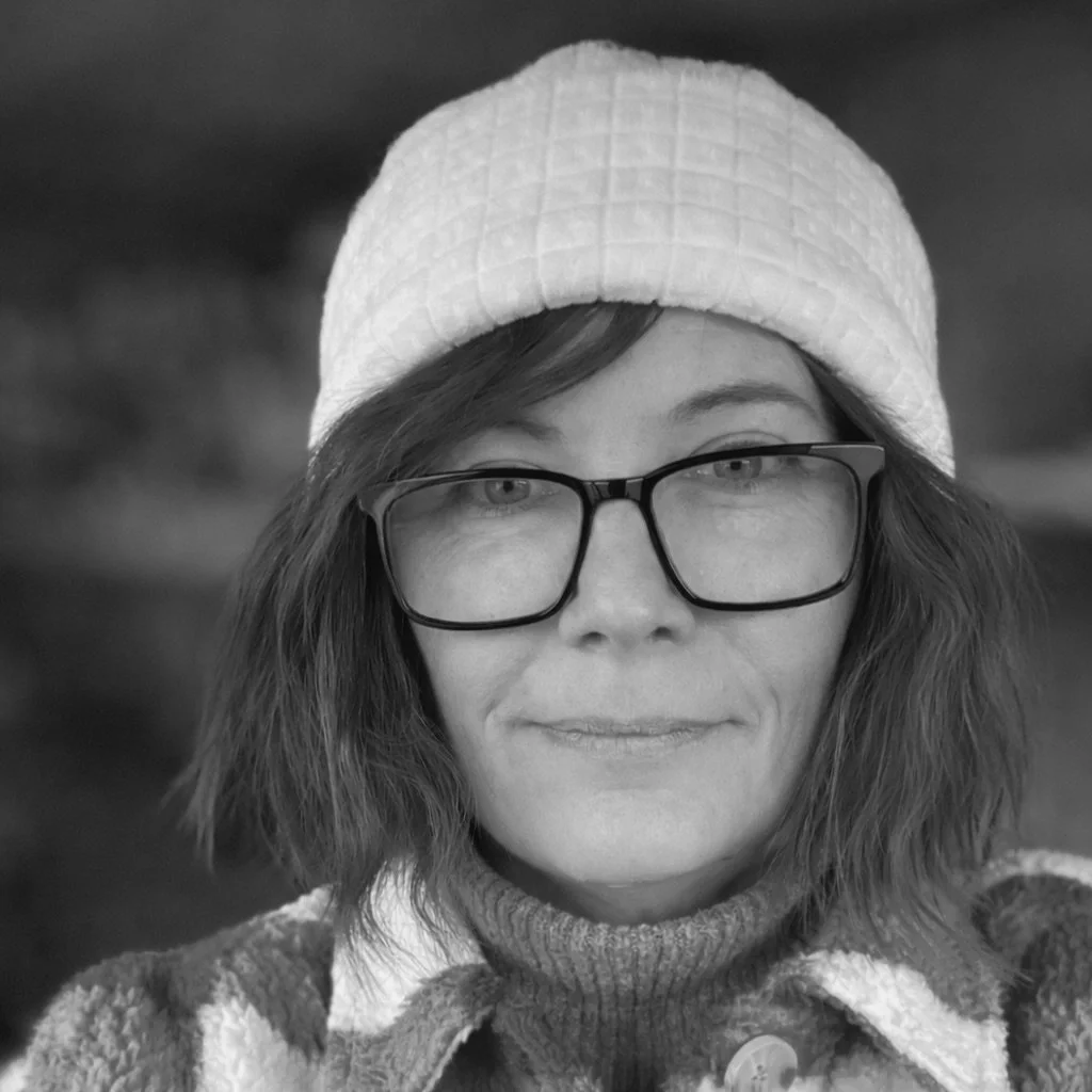 A woman wearing glasses, a quilted white hat, and a knit sweater smiles in an outdoor setting, with blurred natural background.