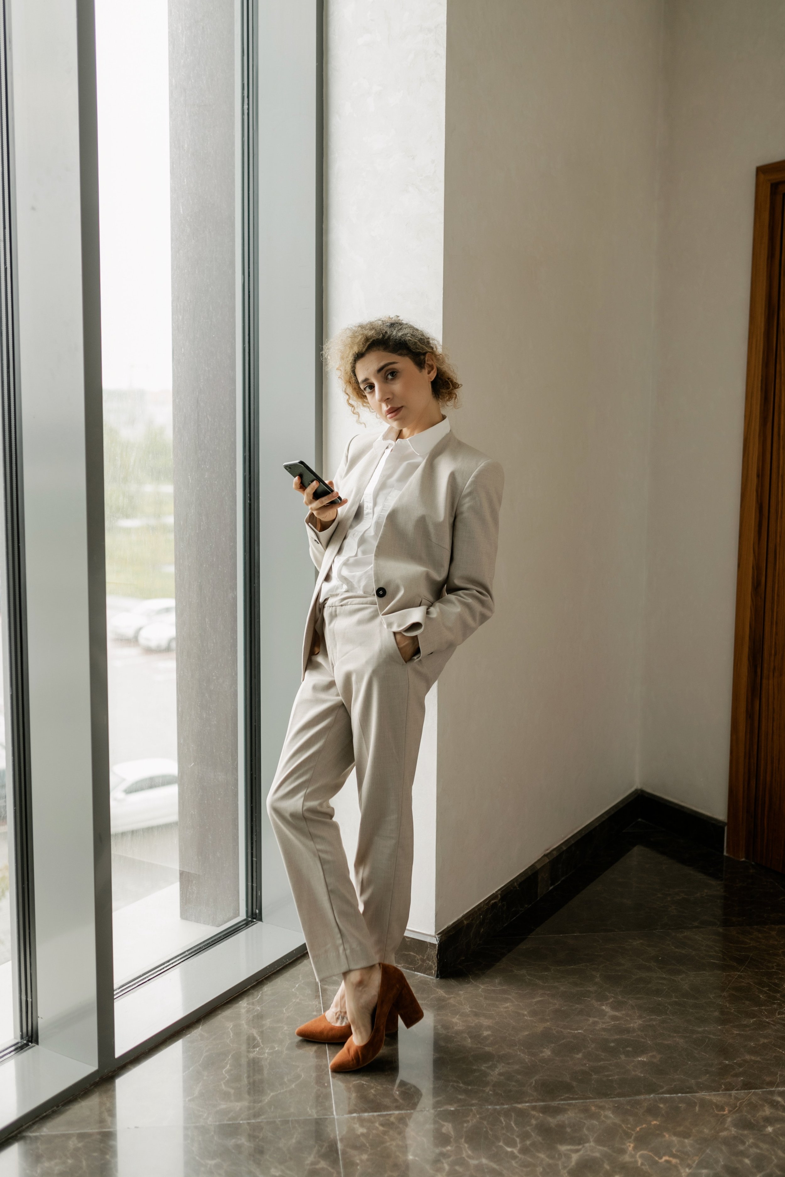 Femme debout dans un couloir près d'une grande fenêtre, portant un costume beige, des chaussures marron, et tenant un téléphone portable. Elle a les cheveux bouclés et regarde vers l'objectif.