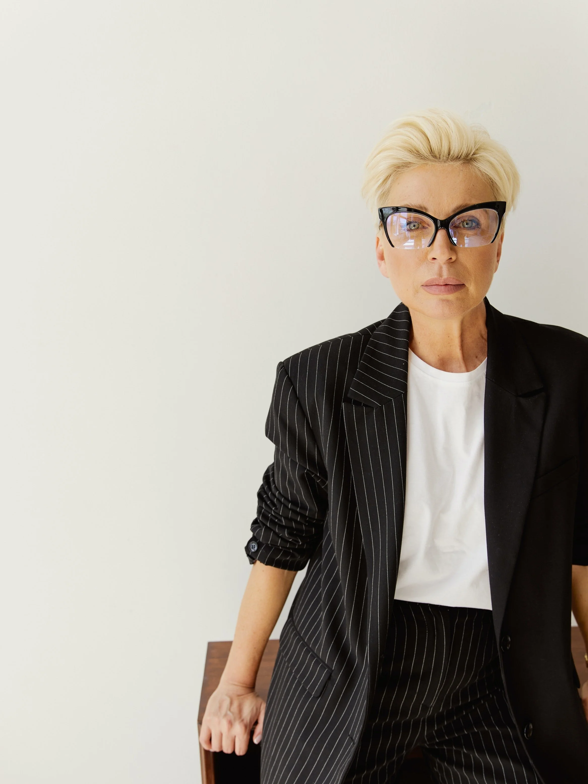 Une femme blonde portant des lunettes noires et une veste noire à rayures et une chemise blanche, assise près d'une table en bois, dans un environnement neutre