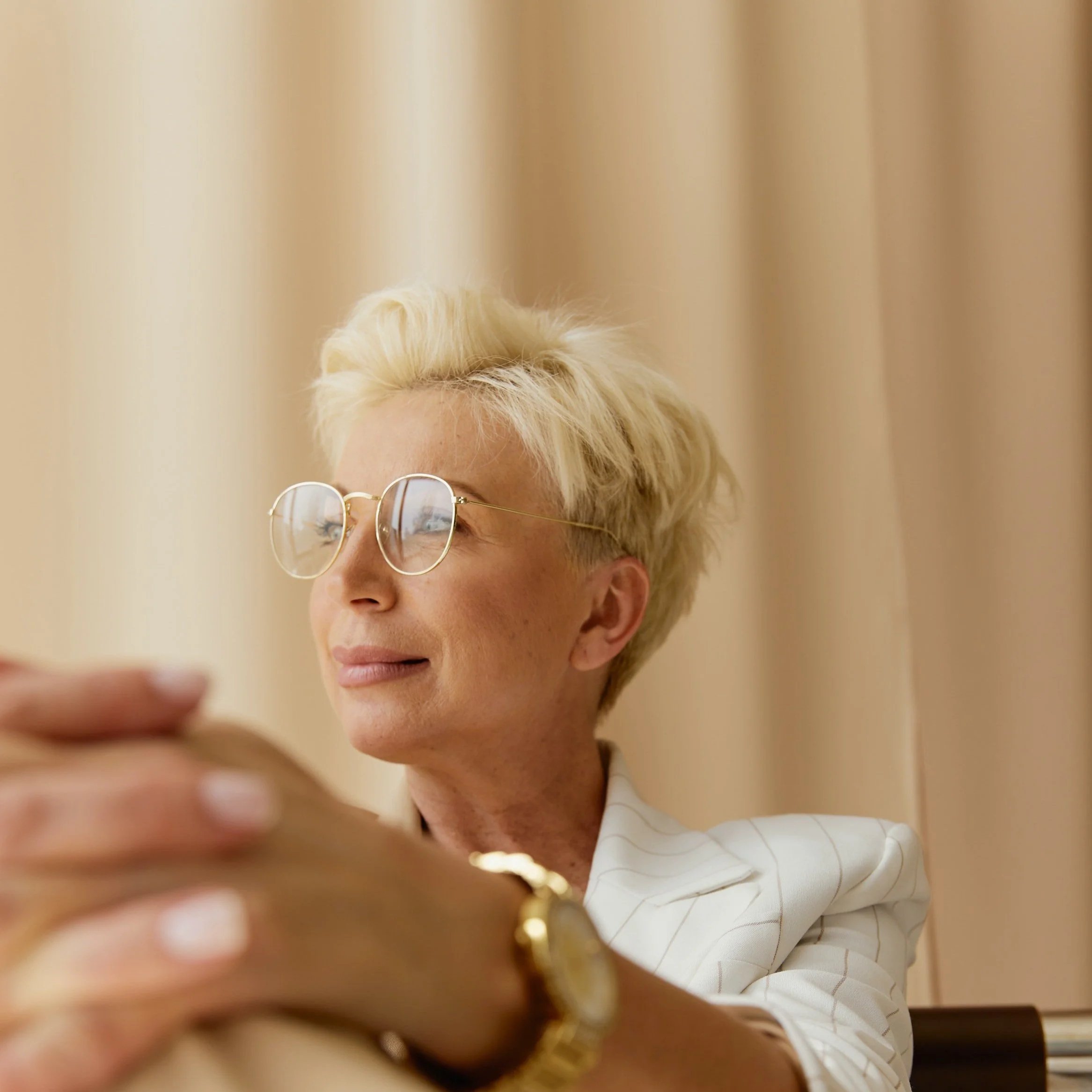 Portrait d'une femme avec des cheveux blonds courts, portant des lunettes rondes avec monture dorée, habillée d'une chemise blanche à rayures dorées, se tenant devant un fond beige, tenant un objet doré ou en or dans sa main