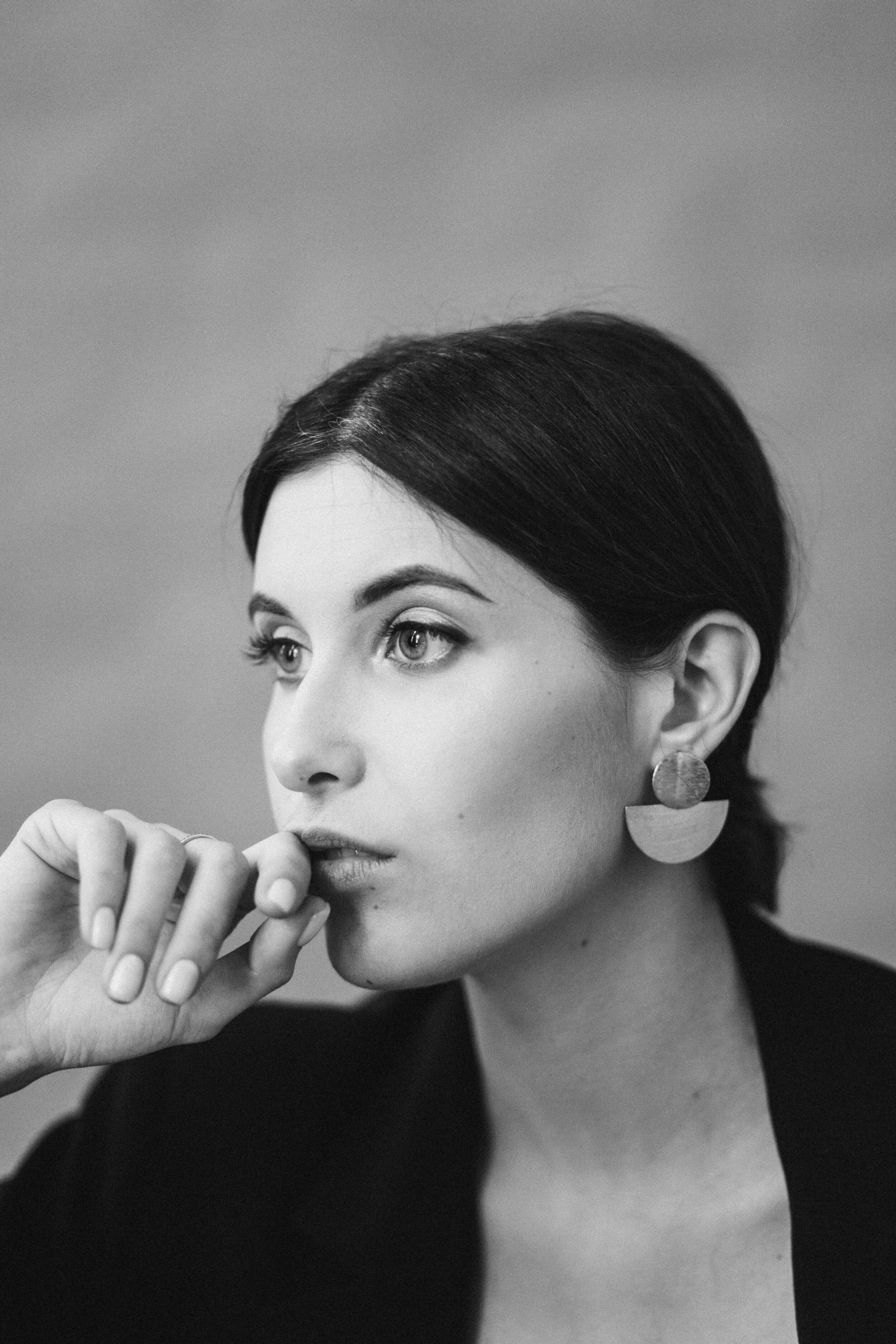 Un portrait en noir et blanc d'une femme avec des cheveux foncés, portant des boucles d'oreilles géométriques, regardant vers la gauche, posant avec sa main près de sa bouche.