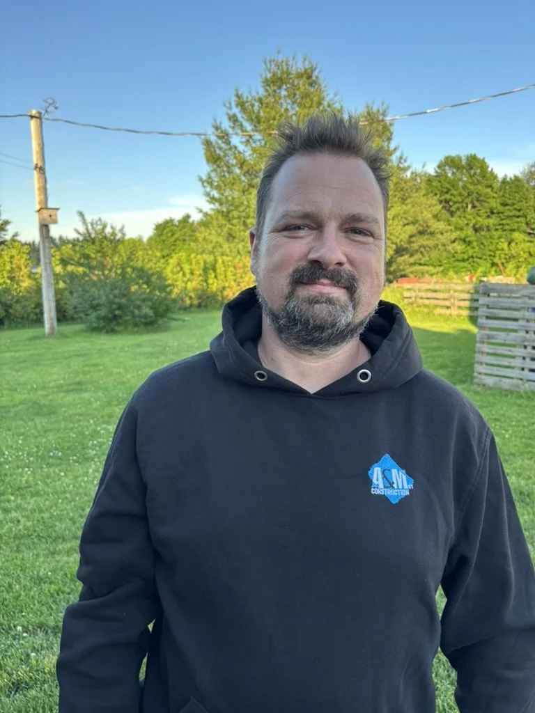 Un homme avec une barbe et une coiffure en épis, portant un sweat noir avec un logo bleu de construction, se tient à l'extérieur dans un espace vert ensoleillé avec des arbres et un ciel bleu clair en arrière-plan.
