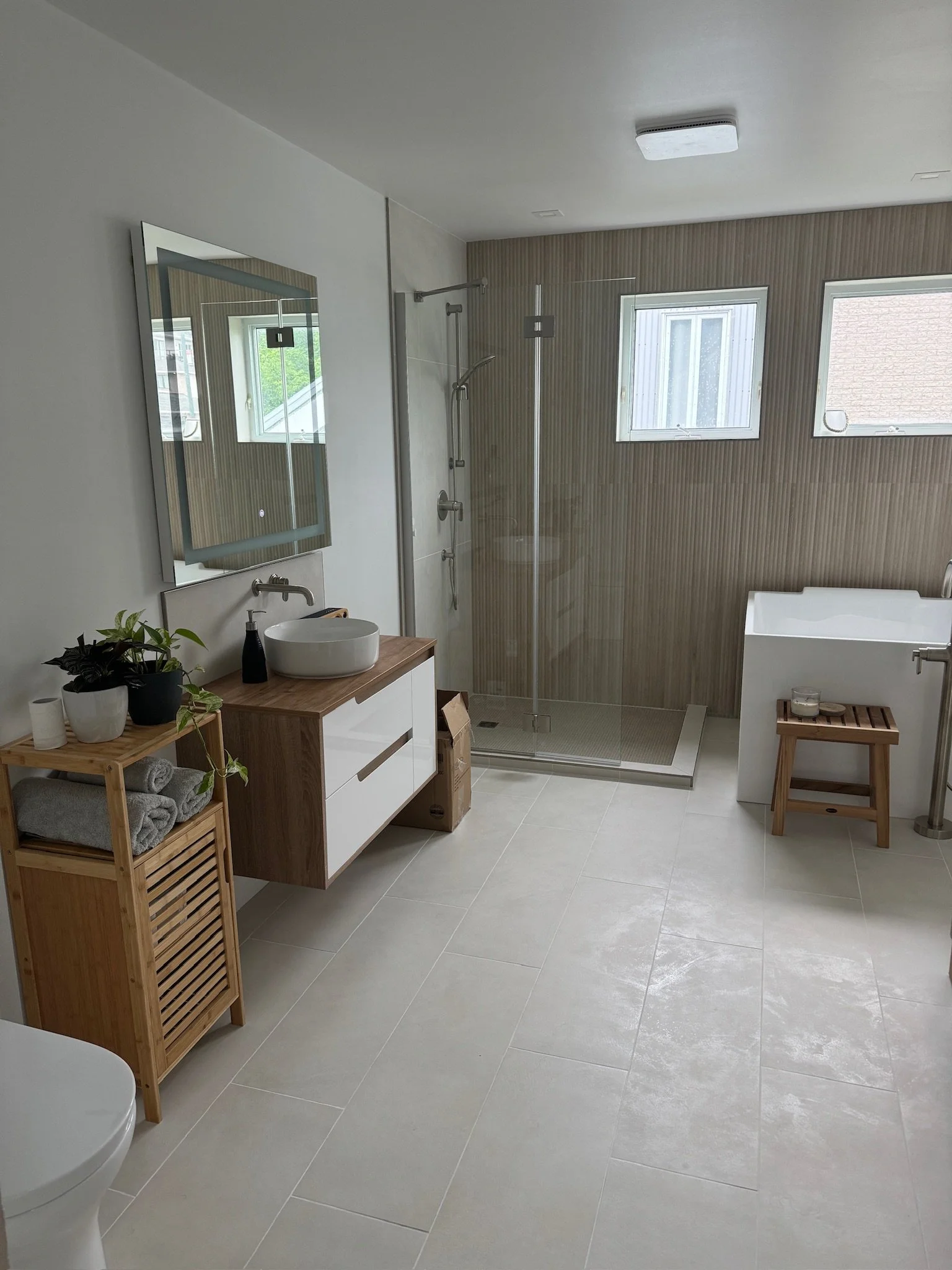 Salle de bain moderne avec douche à l'italienne en verre, deux fenêtres, lavabo sur un meuble en bois clair, miroir rectangulaire, plusieurs plantes, serviettes roulées sur une étagère en bois, baignoire blanche, sol en carrelage beige clair.