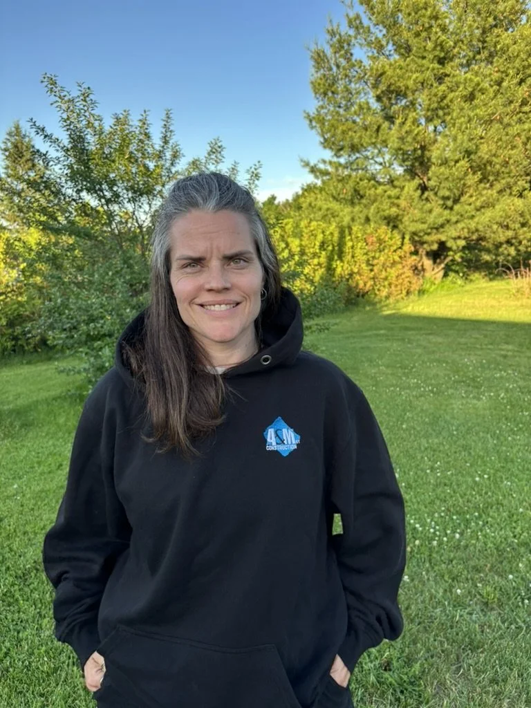 Femme souriante avec des cheveux longs et gris, portant un sweat à capuche noir avec un logo bleu, debout dans un parc lumineux avec arbres verts et un ciel bleu clair.