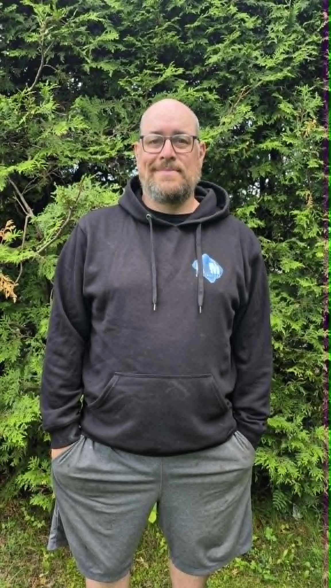 Homme portant des lunettes, pull à capuche noir avec un logo bleu, debout devant un mur de feuillage vert dense.