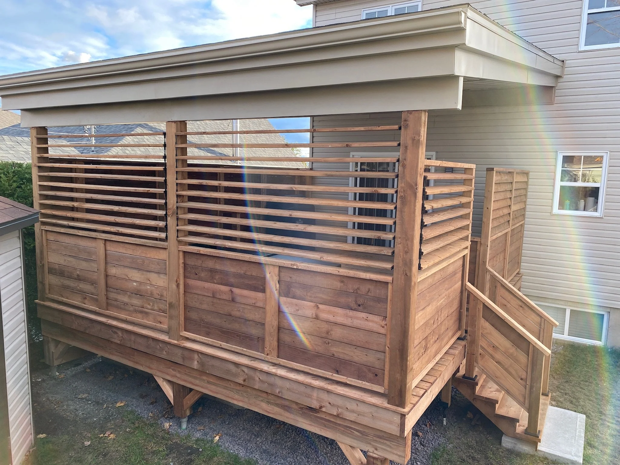 Une nouvelle terrasse en bois en construction attachée à une maison résidentielle, avec des escaliers menant à la zone de la terrasse, sous un ciel partiellement nuageux.