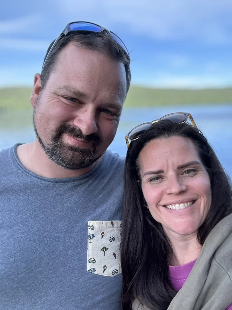 Un homme et une femme souriants prennent une selfie à l'extérieur avec un lac et un ciel bleu en arrière-plan.