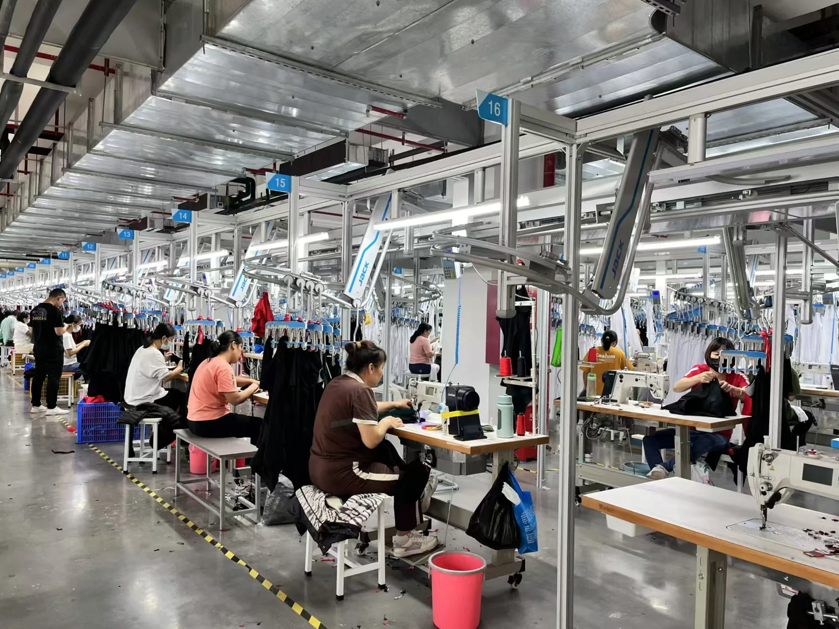 Workers seated at sewing stations in a large, well-lit factory, stitching clothing items with sewing machines.