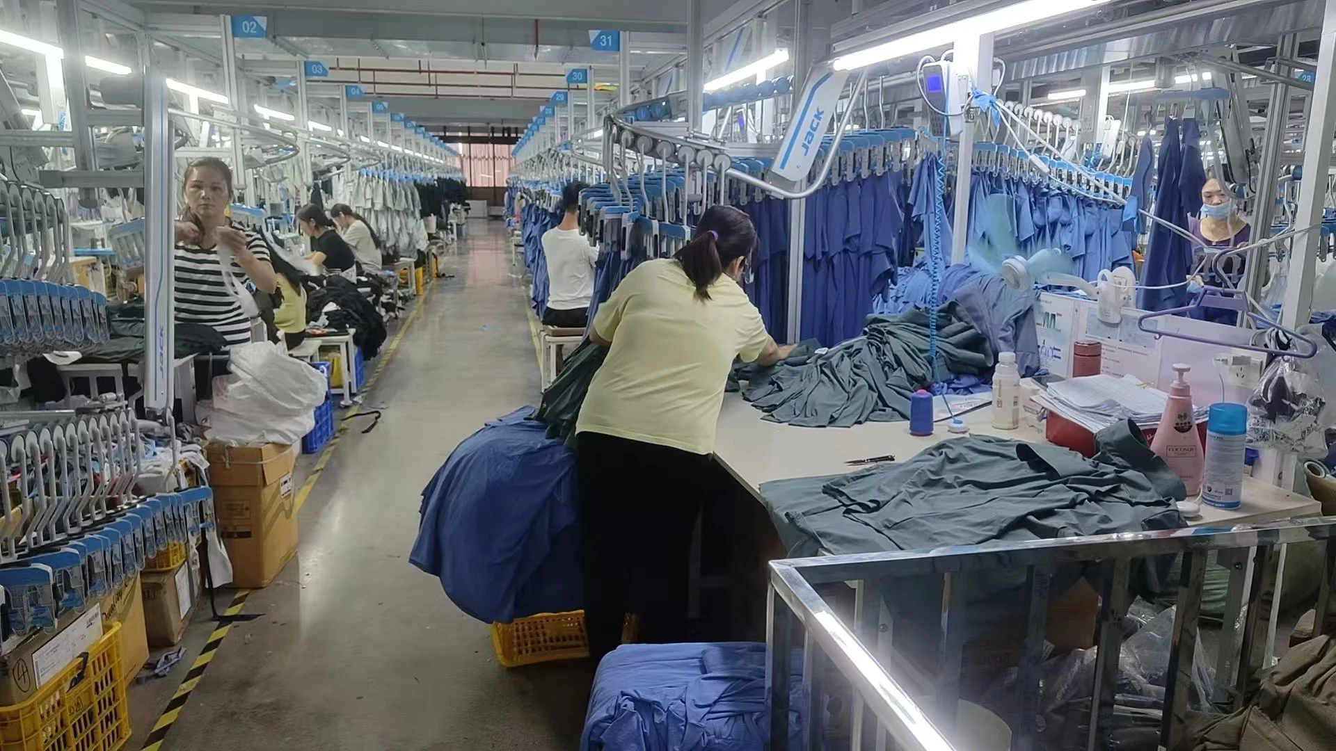 Workers in a factory sewing scrubs with blue fabric in a well-lit workspace equipped with sewing machines and supplies.
