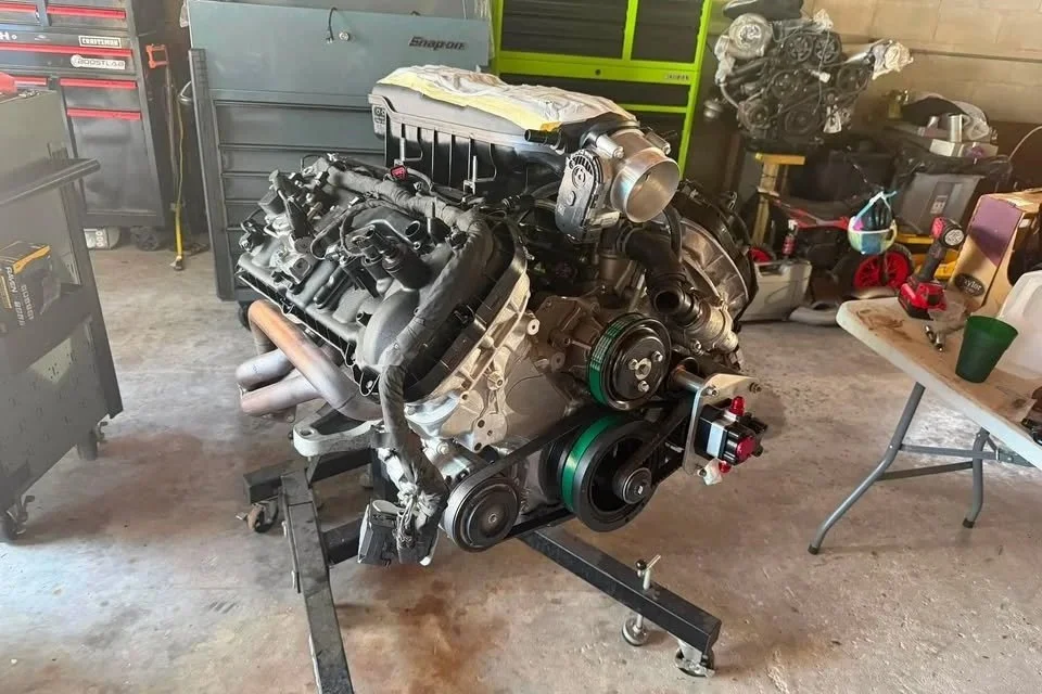 An automotive engine mounted on a stand in a workshop, surrounded by tools and parts.