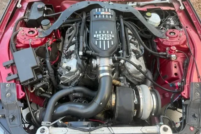 Car engine with a large supercharger, labeled Boss 302, installed in a red vehicle's engine bay.