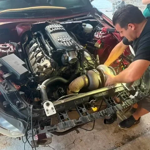 A man working on a car engine, with the hood open, in a garage or workshop.