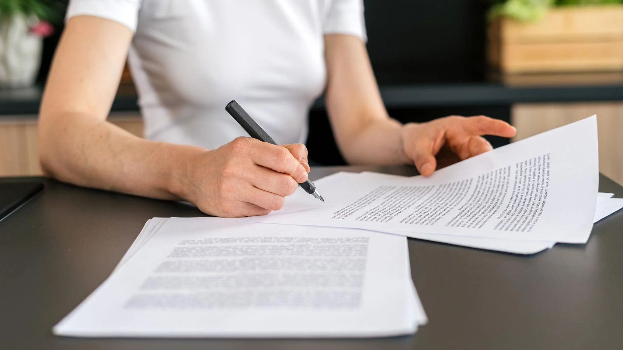 Person signing or reviewing documents on a desk with printed papers, wearing a white shirt.