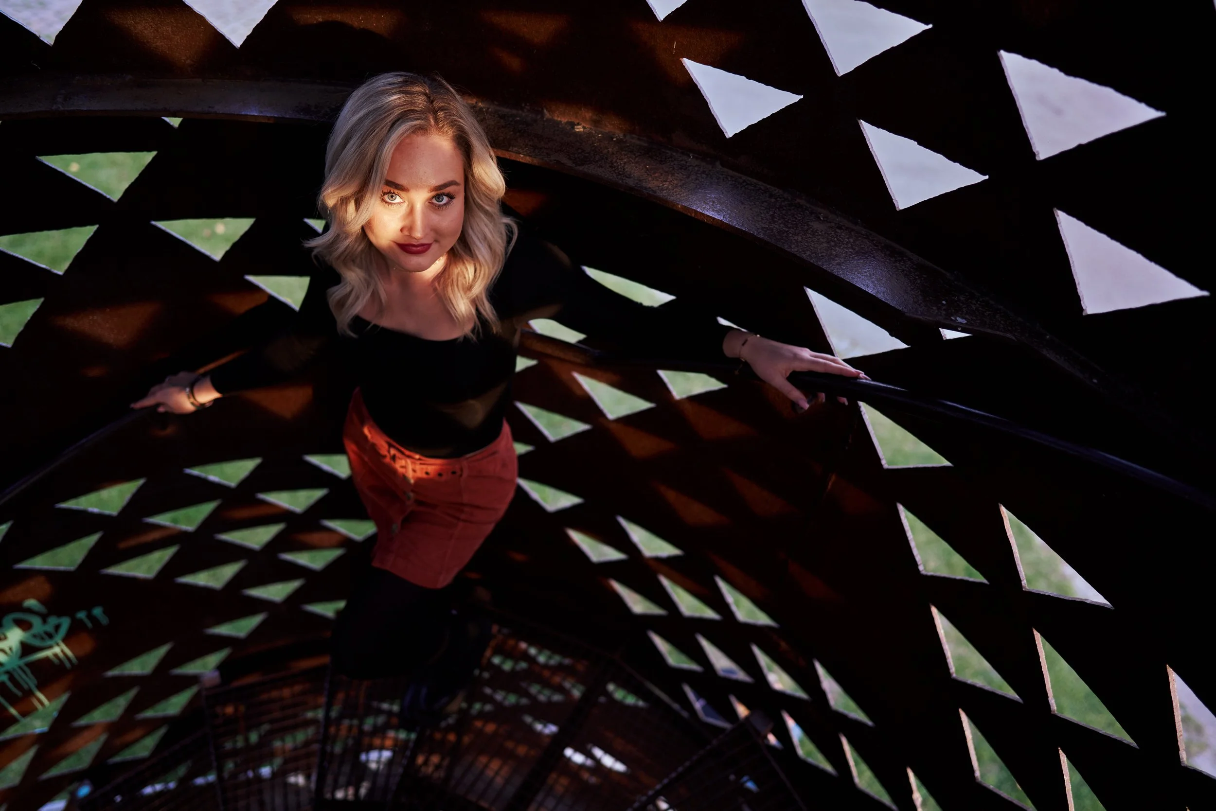 A woman with blonde hair and red lipstick looking up through a tunnel made of rusted metal with diamond-shaped cutouts, with grass visible outside.