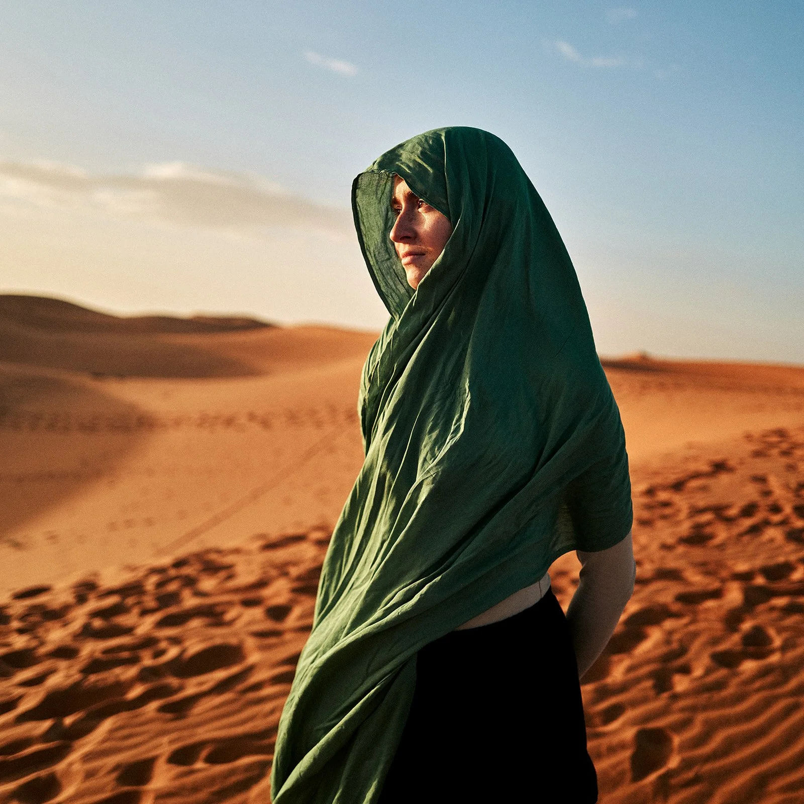 A woman wearing a green headscarf stands in a desert landscape with sand dunes in the background, during sunset or sunrise.