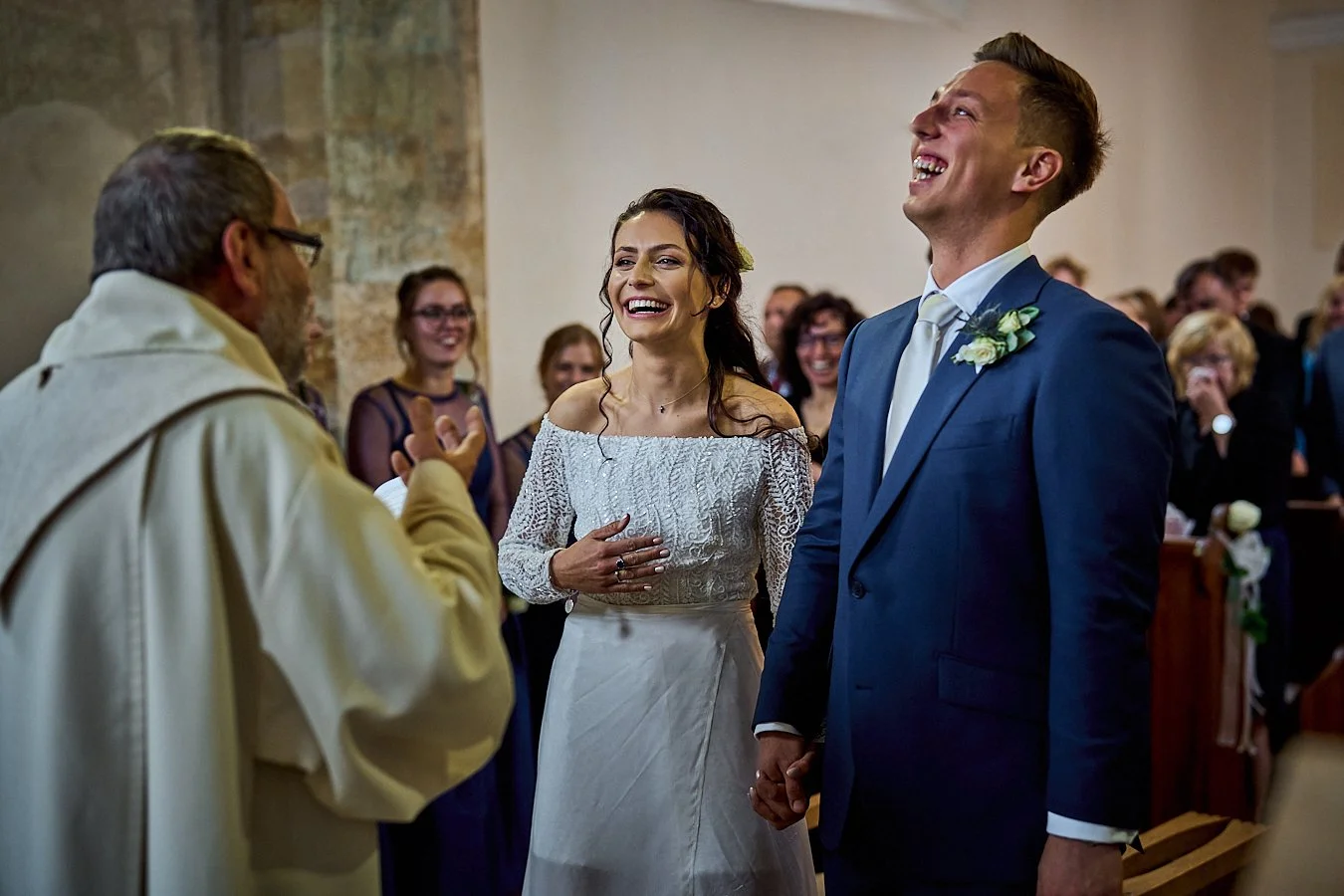 A wedding ceremony with a bride in an off-shoulder white dress and a groom in a blue suit holding hands, exchanging vows or Rings, with a priest or officiant talking to them and guests in the background, smiling and enjoying the moment.