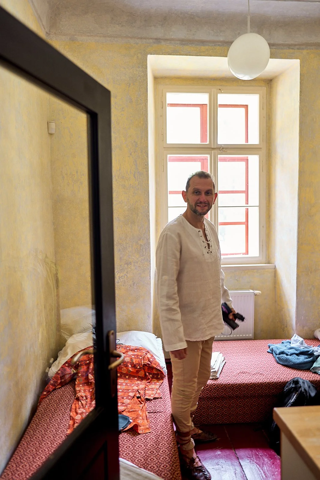 A man with short hair and a beard standing in a small, yellow-walled room, smiling at the camera. The room has a bed with a red-orange patterned blanket, some clothes and a backpack on the bed, a large window with red trim, and a white round ceiling 
