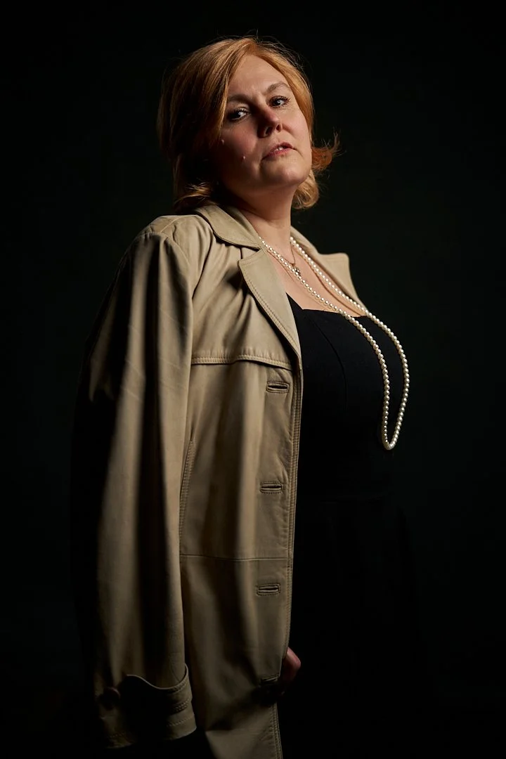 A woman with red hair wearing a beige jacket, black top, and pearl necklaces against a dark background.
